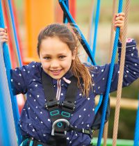 Smiling young person with long brown hair tied back looking directly into the camera whilst crossing the Harlow High Ropes Adventure.