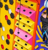 Young person smiling to the camera as they have made it to the top of the orange and blue Clip and Climb wall