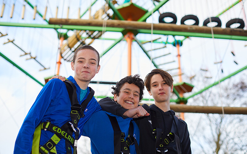 3 smiling teenage boys in front of high ropes course